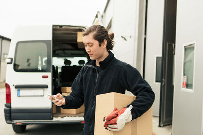 Delivery man using mobile phone while carrying cardboard box against van