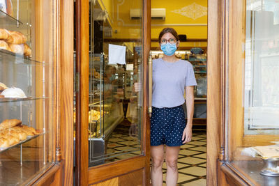 Young female seller in medical mask standing at doorway of cozy bakehouse and looking at camera