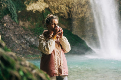 A young woman drinking a hot drink in a mountain canyon in a nature reserve near a large waterfall.