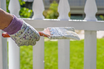 Close-up of hand wearing glove holding paintbrush against fence at back yard