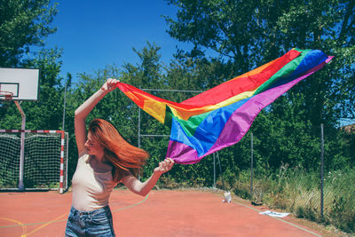 Young woman holding rainbow flag on basketball court against trees