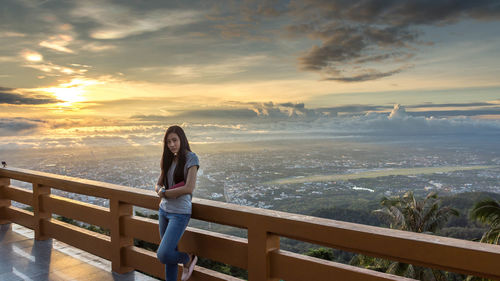 Portrait of young woman standing at railing against sky during sunset