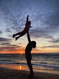 Silhouette man throwing daughter at beach against sky during sunset