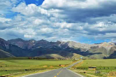 Empty road amidst field and mountains against sky