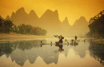 People with birds on boat in lake against mountains during sunset