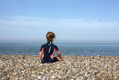 Girl sitting on pebbles at beach against sky