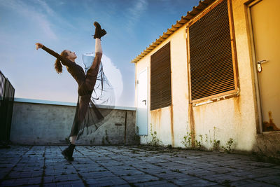 A ballerina in a black skirt  does vertical splits on the roof of a building against the sky
