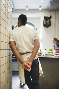 Rear view of male chef standing with hands behind back by wall in kitchen at restaurant