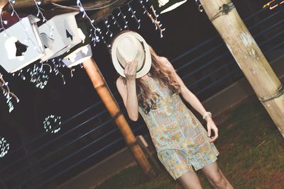 Woman holding hat over face while standing on illuminated field at night