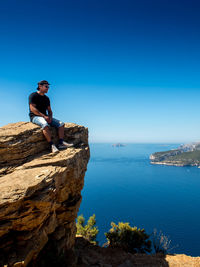 Man sitting on cliff by sea against clear blue sky