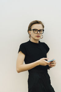 Confident androgynous entrepreneur standing with smart phone against while wall in board room