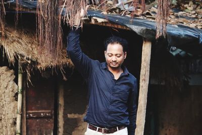 Portrait of young man standing against trees and hut