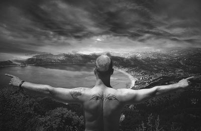 Rear view of shirtless man with arms outstretched against cloudy sky