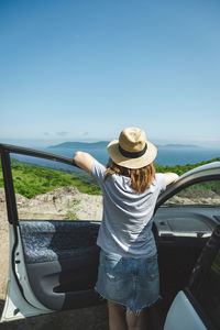 Young woman in hat admires the spacious landscape of sea and islands standing by car. road trips.