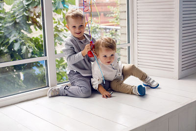 Two brothers sitting by the window in a white studio with green trees on the street