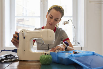 Young tailor using sewing machine while sitting against window at home