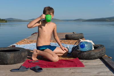 A boy drinks water from a mug while sitting on a wooden pier against lake on a sunny summer day.