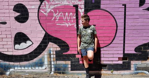 Full length of boy looking away while standing against graffiti wall during sunny day