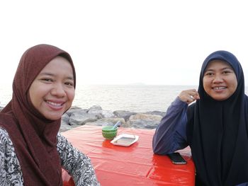 Portrait of smiling female friends in hijab sitting at table against sea