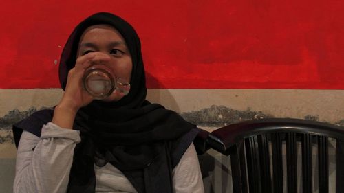 Young woman drinking against red wall