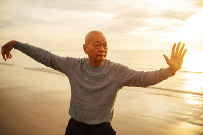 Senior man exercising while standing at beach against sky during sunset