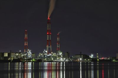 Smoke stacks in illuminated factory by lake against sky at night