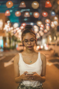 Young woman using phone while standing in illuminated city at night