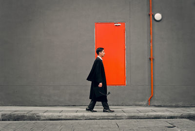 Profile view of young man in graduation gown walking by gray wall