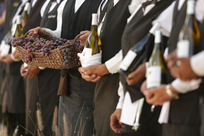 Midsection of people holding grapes and wine bottles on field