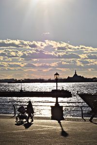 Silhouette woman with baby carriage walking on promenade by sea against sky