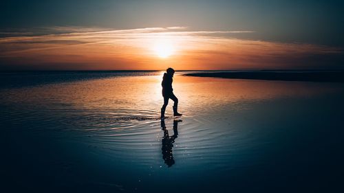 Silhouette boy standing at beach against sky during sunset