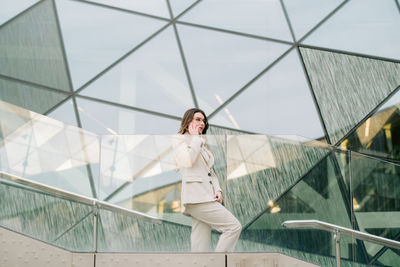 Side view low angle of determined female entrepreneur in elegant suit standing near glass building in downtown and talking on mobile phone while discussing work issues and looking away