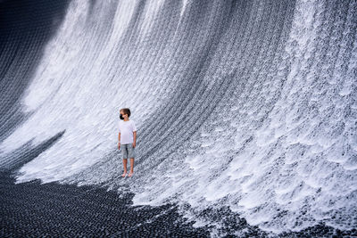 A boy in denim shorts and a white t-shirt looks at an artificial waterfall. 