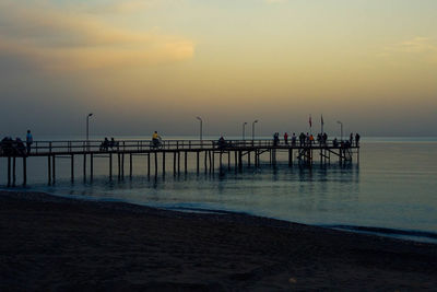People on pier at beach against sky during sunset