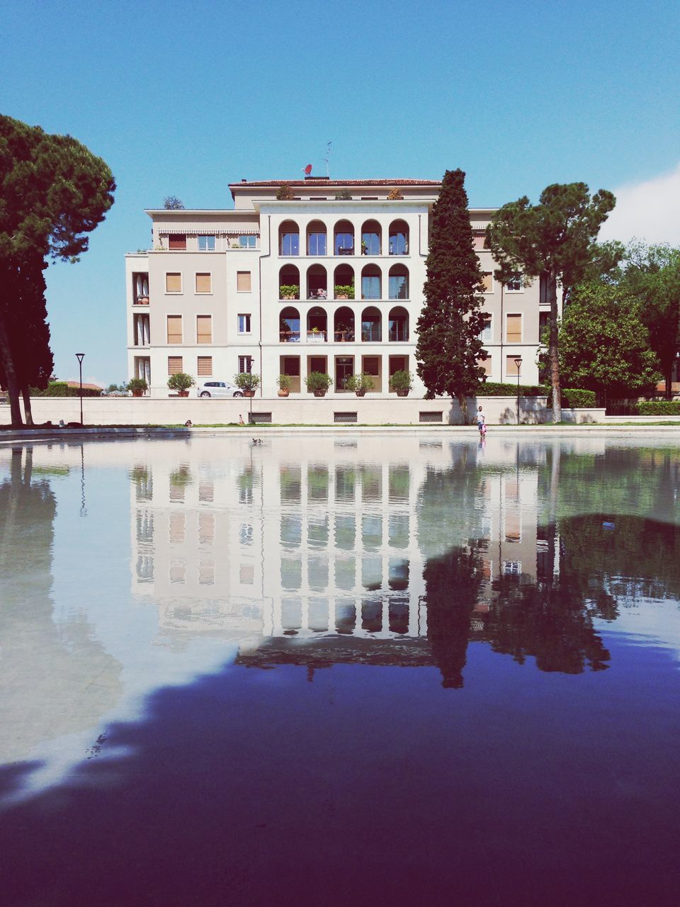 architecture, built structure, building exterior, water, reflection, waterfront, tree, clear sky, fountain, sky, pond, architectural column, day, blue, famous place, sunlight, outdoors, city, travel destinations, building