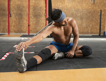 Muscular male athlete in stylish sports clothes stretching legs and back on floor in gymnasium white looking away