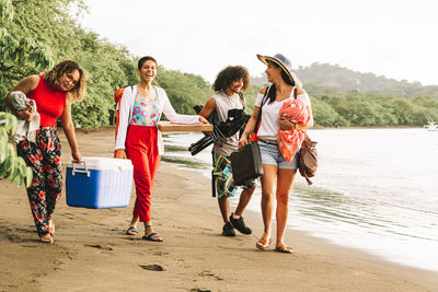 Full body of cheerful young friends carrying food and supplies for summer picnic on beach of costa rica