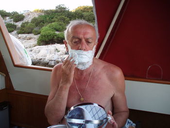 Shirtless senior man applying shaving cream on face while sitting in boat