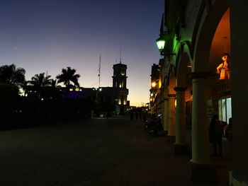 Illuminated street amidst buildings against sky at dusk