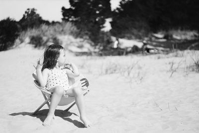 Cute girl sitting on chair at sandy beach during sunny day