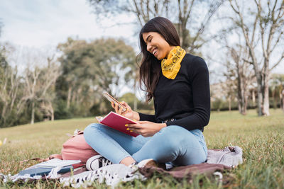 Young ethnic female university student in denim outfit browsing on smartphone while reading book in park