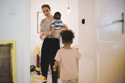 Mother carrying toddler looking at son while standing in domestic room