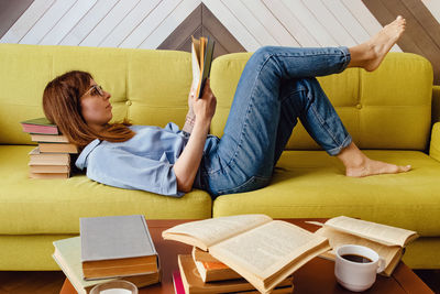 Young woman reading on the sofa at the living room with a lot of closed and opened books around. 