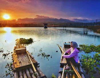 Woman sitting on wooden raft in lake against sky during sunset