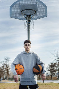Serious hispanic sportsman with basketballs looking at camera while standing near basketball hoop on sports ground during training on street