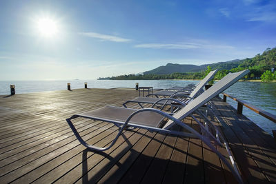 Empty chairs by swimming pool by sea against sky on sunny day