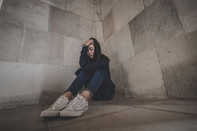 Young woman looking away while sitting on floor against wall