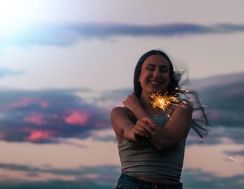 Smiling woman holding lit sparkler against sky during sunset