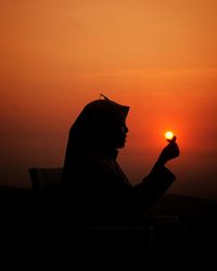 Silhouette woman sitting like hold and touch the sun against orange sky