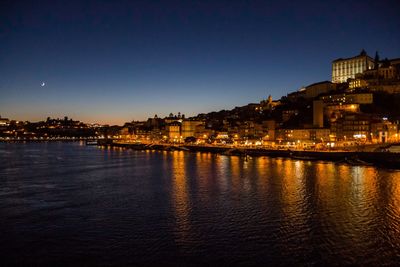 Illuminated buildings by river against sky at dusk